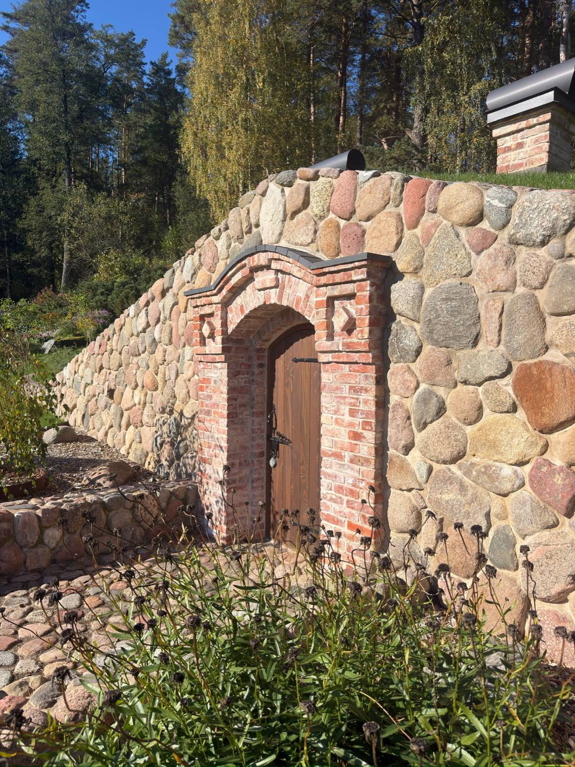 Lång mur av rundad natursten i olika kulörer, med en portal i återbrukat tegel och murade skorstenar ovanför